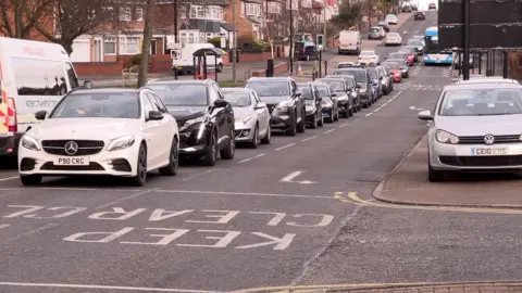 A row of cars waiting for a junction to be clear of other vehicles, while one of the carriageways, currently not a dedicated bus lane, is occupied by a blue and white bus.