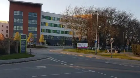 The front of Pinderfields Hospital, viewed from a road. It is a modern hospital building. Bare trees, roadsigns and signposts are all in front of it.