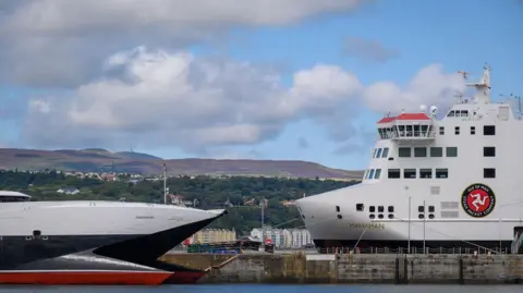 Manx Scenes The Manxman ferry docked in Douglas Harbour next the Manannan. The ferries are white, black and red. There are green hills in the background and a blue sky with white clouds.