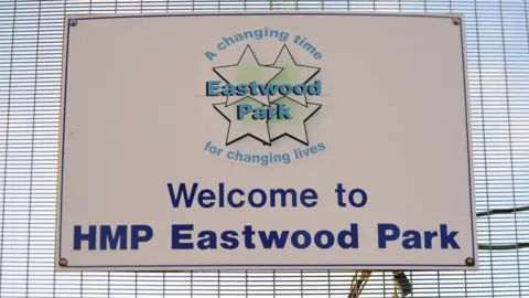 A sign on metal fencing reads 'Welcome to HMP Eastwood Park' alongside the prison's logo which is made up of four stars.