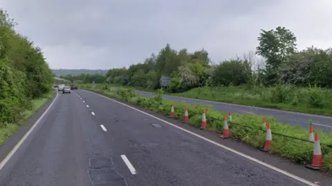 Google A dual carriageway with a few potholes visible and a line of orange cones in the central reservation. There are trees and bushes adjacent to the road.