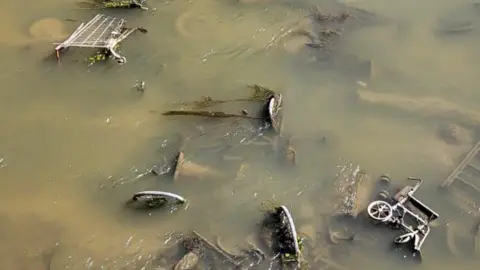 Ben Bishop Rubbish items in the River Parrett. A shopping trolley and a tricycle are visible in the water, which is a muddy brown colour
