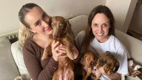 Dog breeder Sophie Mortimer has blonde hair tied back in a ponytail, she is wearing a brown top and holding up a caramel-coloured long-haired dachshund puppy. To the right of her Bethan Lee has brown long hair, she is wearing a white T-shirt and holding two small caramel-coloured long-haired dachshund puppies.