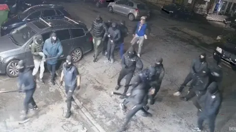 Several people stood in a group, many are wearing balaclavas. Some of the group can be seen holding weapons in their hands. In the background are parked cars. 