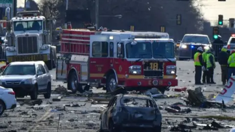 Crash debris on the road while a burned out car is visible in the foreground. You can also see emergency service personnel in hi viz jackets and fire truck in the background. The road looks to be cordoned off. 