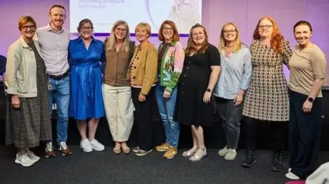 NHS North Bristol Hospitals Trust The team at Southmead hospital is lined up for a picture at an official OdonAssist launch event in London. They are standing on a stage and smiling at the camera.