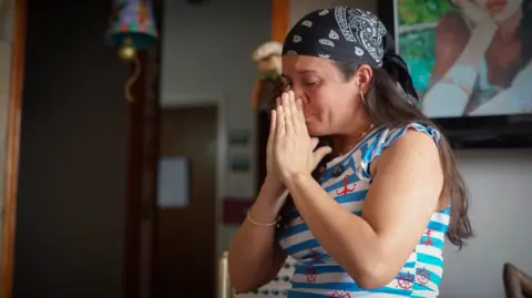 Adriana Briceño hold her hands in front of her face as she fights back tears, while sat in a the living room of her home