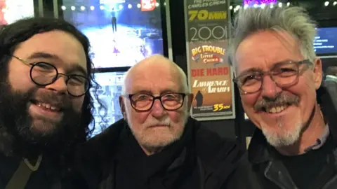 Jon Spira Spira and Hodges with Rhys Jones in a selfie taken in a cinema foyer. John has long dark hair, a dark beard, and black rimmed glasses. Rhys Jones has grey hair, a grey goatee, and grey-rimmed glasses.