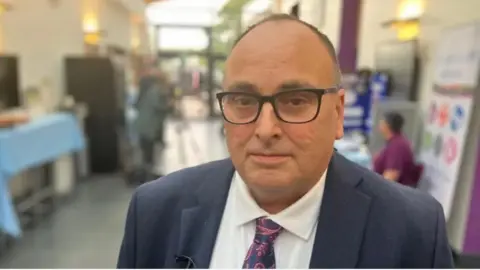 A man in a suit, paisley tie and black-rimmed glasses.