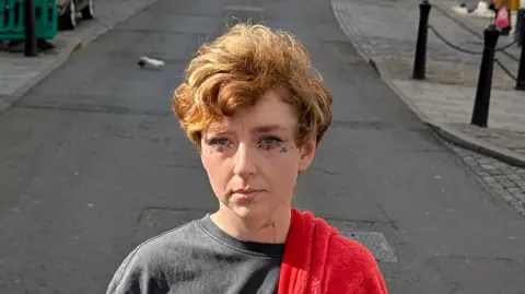 Ed Gold/Positive Futures Erin wearing a grey T-shirt with a red blanket draped over her left shoulder. She has short fair hair and is standing in the middle of a road. She has small tattoos beneath her eyes.