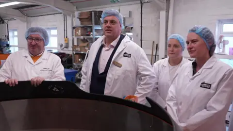 Four people in white coats and food hygiene hair nets stand around a large machine that roasts coffee beans.
