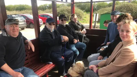 James Hewett Passengers sit in a train carriage which has open windows which are framed with wood.