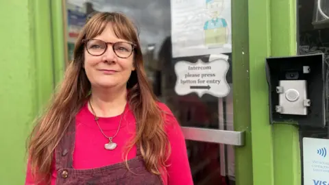 Simon Thake/BBC A woman in a bright pink long sleeved top stands in front of a green shop front and intercom system