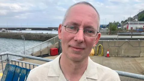 William is looking at the camera. He has balding grey hair and blue eyes. He has thin glasses on and a metal chain. He is wearing a linen shirt. Behind him is a ramp and a bay.