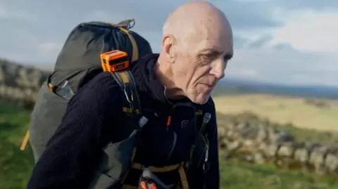 The Montane Spine Race Runner Kevin Munt is running along the Pennine Way with a grey backpack on. He is wearing a navy fleece. He is running the Spine Race ultramarathon and looks tired in his facial expression.