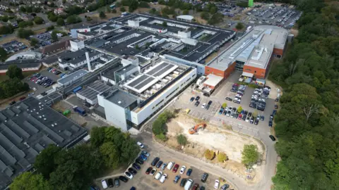 Steve Hubbard/BBC An aerial view of a hospital which has several different buildings and car parks located at the top and bottom of the site. There are rows of trees on the right.