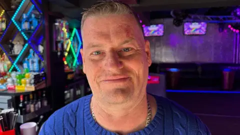 A man in a blue cable knit jumper with a heavy silver chain and short fair hair smiles. Behind him a club can be seen with neon lighting around seating areas and behind the bar.