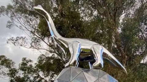 A steel dinosaur sculpture stands on top of a geometric ball in New Zealand.