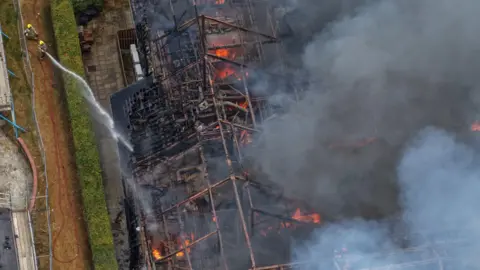 Ian/Yorkshire Visions Media An aerial view of a large building engulfed in flames, with its roof collapsed and thick smoke rising. Two firefighters on the ground spray water from a hose toward the burning structure, surrounded by a paved walkway and hedges.