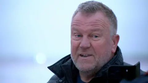 Barry Elliott has short greying hair, combed forwards from the temples, with a slight peak at the forehead and a grizzled white grey beard and slight stubble moustache. He is wearing a black coat with the collar up. He is mid sentence, with his mouth open and forehead wrinkled and looks slightly annoyed.