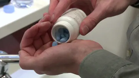 File image showing a man tipping a small white plastic bottle containing blue PrEP pills until one falls into his hand. A bathroom sink and blue bottle of water can be seen in the background 