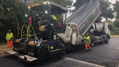 Oxfordshire County Council Close-up view of freshly laid asphalt on a rural roadway. A road resurfacing team in high-visibility orange safety clothing operates paving equipment under an overcast sky, with fields stretching alongside the road.