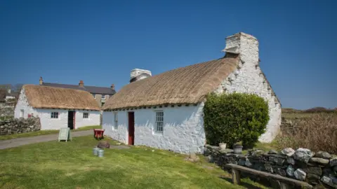 MANX SCENES The exterior of the cottage before the works, it is a white straw thatched building, there is another similar building perpendicular to it.