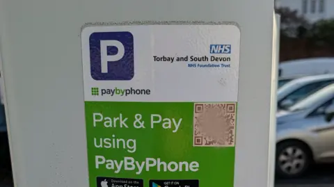 Emma Bovey The front of a Park and Pay machine at Totnes hospital with a square QR code image in the top right corner and cars parked behind.