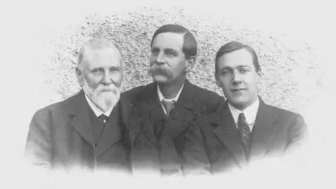 Chris Stretton A black and white photograph showing three men wearing dark coloured suits all stood next to each other. The man on the left has a long white beard, while the man in the middle has a thick moustache and the man on the right is clean shaven with his hair swept to one side.