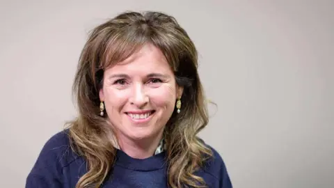 Mary's hair is brown with highlights, and is short at the front and falls in loose curls over her shoulders. She is wearing yellow earrings and a navy round neck jumper. 