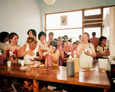 Martin Parr/Magnum Photos A group of children and adults in a crowd waiting for hot dogs behind a wooden counter, most are in swimwear