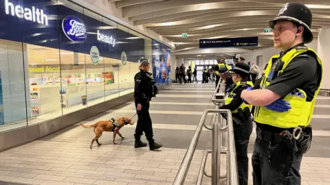 The inside of Birmingham New Street station with a Boots pharmacy on the left with glass windows. A policewoman with black uniform labelled for the British Transport Police has a brown dog on a lead. On the right, behind silver-coloured barriers, there are several police officers with yellow high-visibility vests, black radios, black police hats and blue gloves.