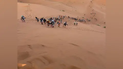 Marc Lockwood Marc Lockwood pictured among a number of participants in the Marathon des Sables in the Sahara Desert. A number of small participants can be seen walking up a sandy hill.