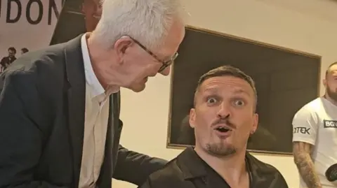 Paul Gilfeather Frank Gilfeather hablando con el campeón de peso pesado Oleksandr Usyk, Frank viste un traje negro con una camisa blanca riendo y Usyk viste una camisa negra mientras mira a la cámara sorprendido.