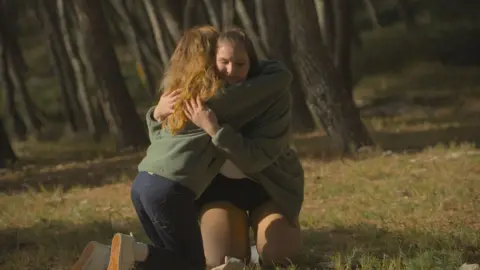 Channel 4 Katie is kneeling on the ground and another woman is hugging her. They are both wearing green jumpers and are in a woodland area.