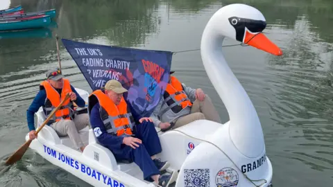 A swan-shaped pedalo with four people on board sets off on the River Thames in Lechlade. The two men at the front are pedalling while the crew members at the back row. There is a large banner between the crew for Combat Stress, a mental health support charity for veterans. All members of the crew are wearing bright orange life jackets. 
