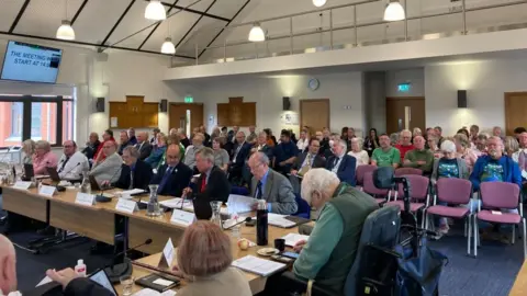 Laura Coffey/BBC Council chamber with pitched roof and small half-egg lampshades hanging from ceiling. There is a mezzanine floor at the back with railings. Councillors and members of the public are sitting in purple seats on the ground floor. The councillors are sitting behind wooden desks and have papers in front of them.
