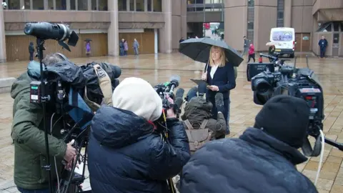 PA Media CPS prosecutor Ursula Doyle delivers a statement to camera crews outside Liverpool Crown Court. She is holding a clipboard and sheltering under an umbrella. 