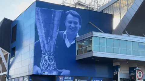 A painted image of Sir Bobby Robson holding a trophy on the side of Portman Road stadium. The image is painted in blue and white.