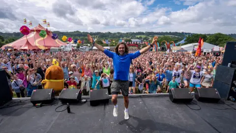Joe Wicks, wearing a blue workout top and cargo shorts, stands on a stage with his back to the crowd holding his arm in the air to pose for a picture. He is jumping slightly off the ground. Thousands of people can be seen in the background, many who are cheering with their raised to the skies. A red circus tent can be seen in the background as well as colourful festival flags on the horizon.