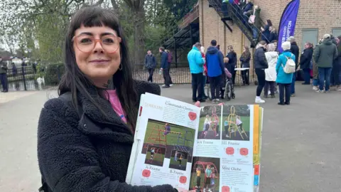 Kate Bradbrook/BBC Amber McAllister has dark hair which is straight and worn down. She is looking directly at the camera and is smiling and is wearing a pink T-shirt an black coat. She is holding a brochure from the 1990s with photographs in of when she modelled for pictures taken on playing equipment at the park. Behind her is a queue of people waiting to go on the water chute. There is also a purple sign with white writing on it which says, BBC Radio Northampton.