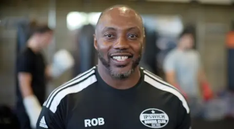 Jamie Niblock/BBC Rob smiles at the camera wearing a black adidas t-shirt with his name printed in white, as well as Ipswich Boxing Club. He has a smart goatee beard,