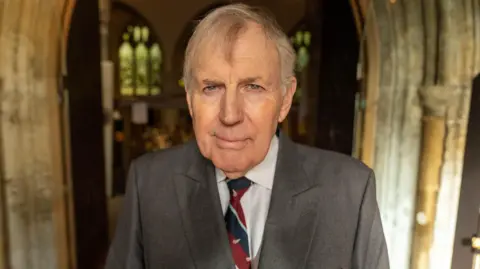 Ian Palmer/ BBC A man with grey hair looks at the camera. He wears a grey suit and white shirt with an navy and burgundy diagonally striped RAF tie