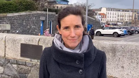 Lindsay De Sausmarez looking at the camera. She has dark hair, is wearing a light grey scarf and a dark grey coat.