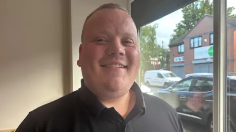 Simon Thake/BBC A jolly looking man in a black polo shirt and with short cropped hair smiles at the camera.