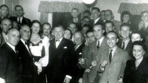RAFA Jersey A black and white indoor photo of a group of predominantly men with a few women. The men are wearing suits and ties and the women are formally dressed. The people are crowded together. Behind them are two windows with pelmets and curtains.