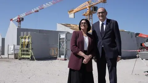 Home Secretary Shabana Mahmood with French Interior Minister, Laurent Nunez, on the site of a new detention centre that is being built in Dunkirk, France. Behind them there are partially constructed buildings and cranes.