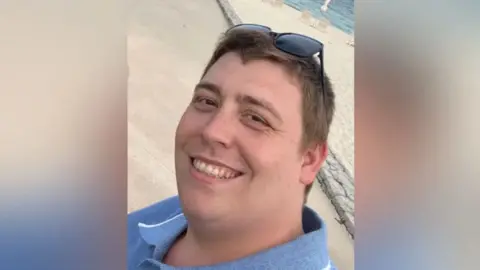 Family photo Mathew Chapman wearing a blue collared shirt and smiling at the camera, he is standing next to the beach and the sea is visible behind him