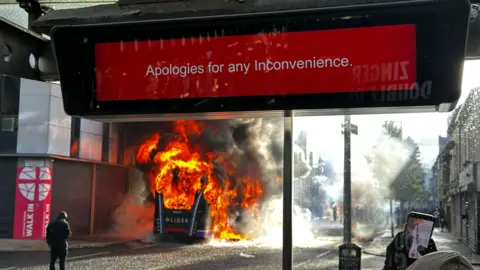 A Glider bus on fire in front of a bus sign which says 'Apologies for any inconvenience'.