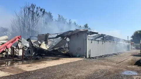 Suffolk Fire and Rescue Service A large storage building which has been involved in a fire. Smoke is billowing out the building. The roof has been burnt and destroyed in the blaze.